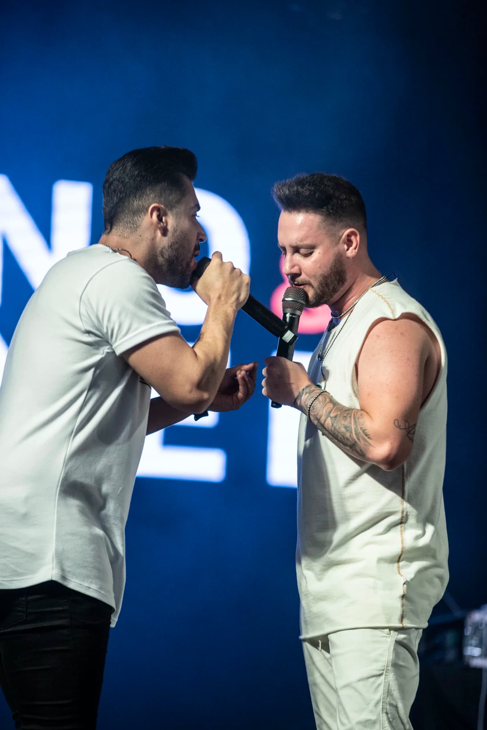 Bruno e Denner cantando frente a frente sob luz azul